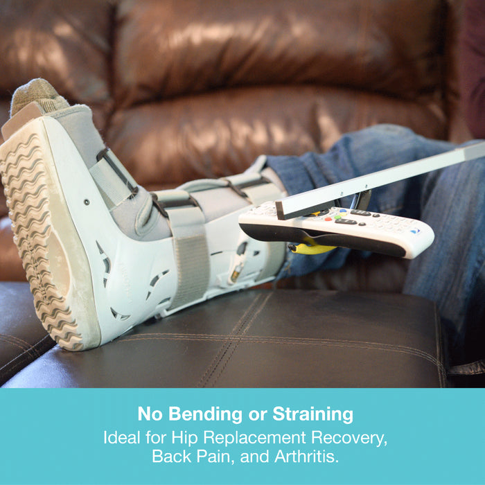 A white and gray leg cast boot with a white remote control resting on it, shown on a person's leg in a brown leather recliner. A teal banner at the bottom contains text reading 'No Bending or Straining' and 'Ideal for Hip Replacement Recovery, Back Pain, and Arthritis.