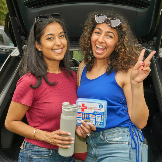 Two women standing in a car trunk with a first aid kit and water bottle.

