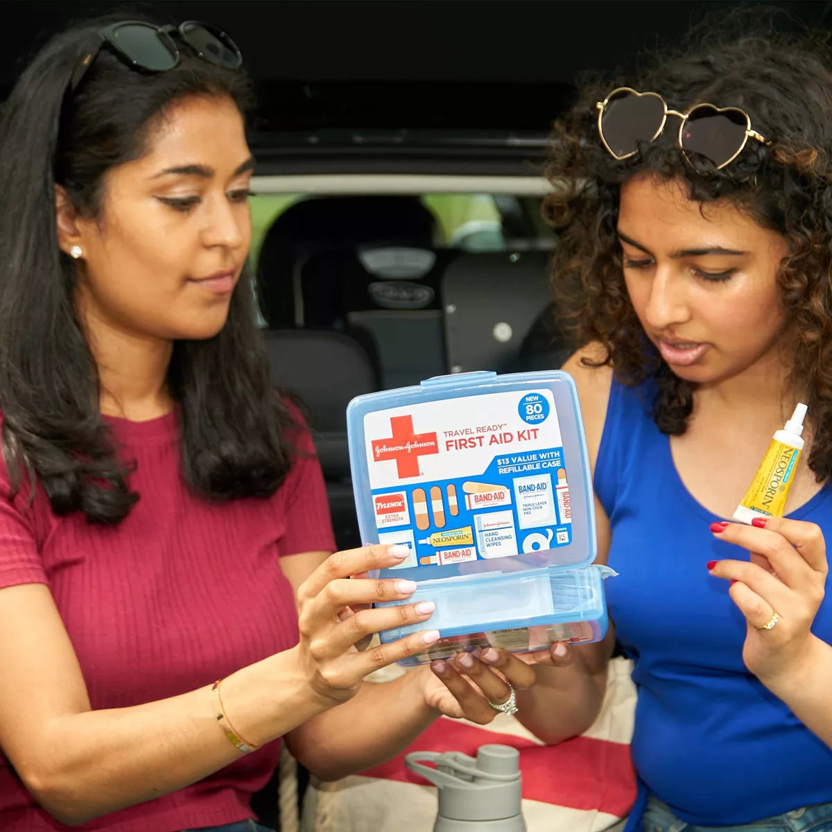 Two women examining a first aid kit in front of an open car trunk.

