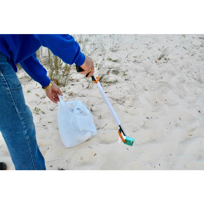 A person in a blue sweatshirt and jeans uses a long-handled white and orange beach pick-up tool to collect trash from white sand next to a white plastic bag.