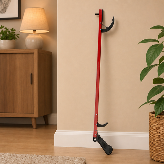 A red and black reacher/grabber with a long handle and curved hook-like end, leaning against a beige wall in a styled indoor room with a lamp, cabinet, plant, and potted plant.