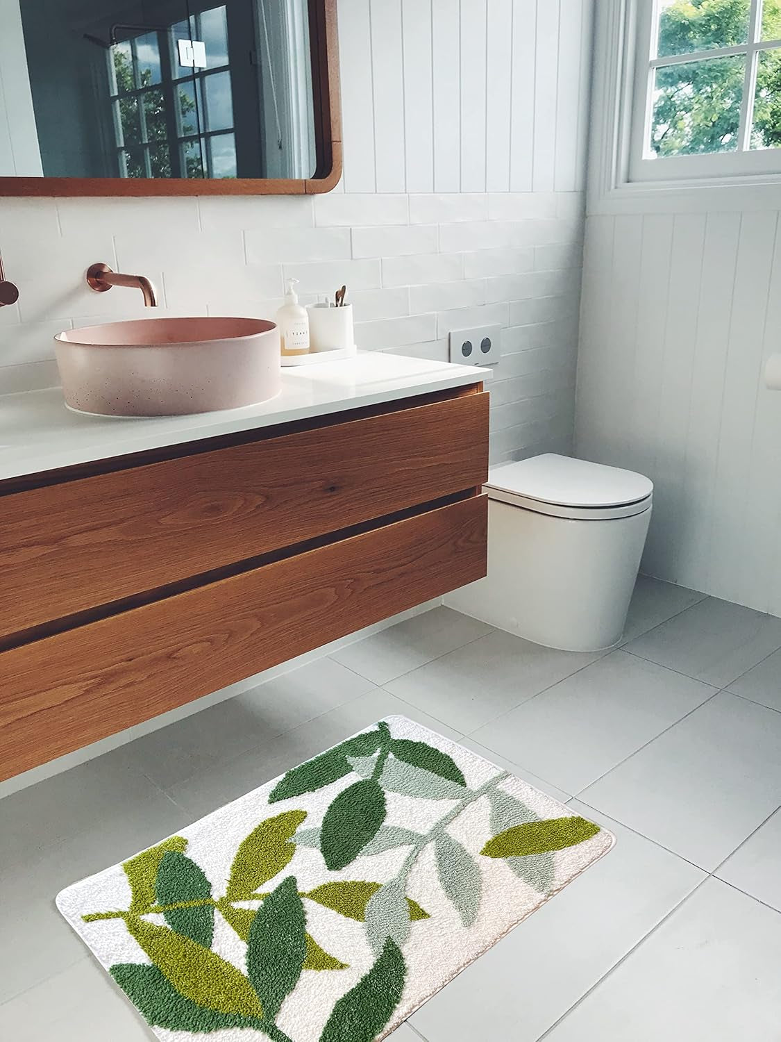 Bathroom with wooden vanity, pink sink, and white toilet with a green leaf-patterned rug on the floor.


