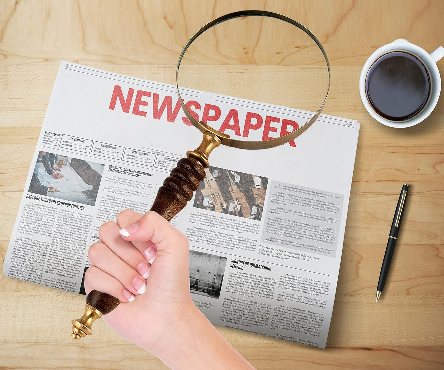 Hand holding a magnifying glass over a newspaper on a wooden table with a cup of coffee and pen.